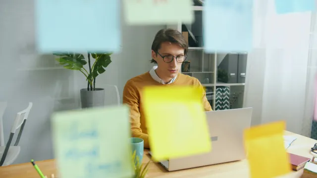 A man working on a laptop behind a glass wall covered in sticky notes